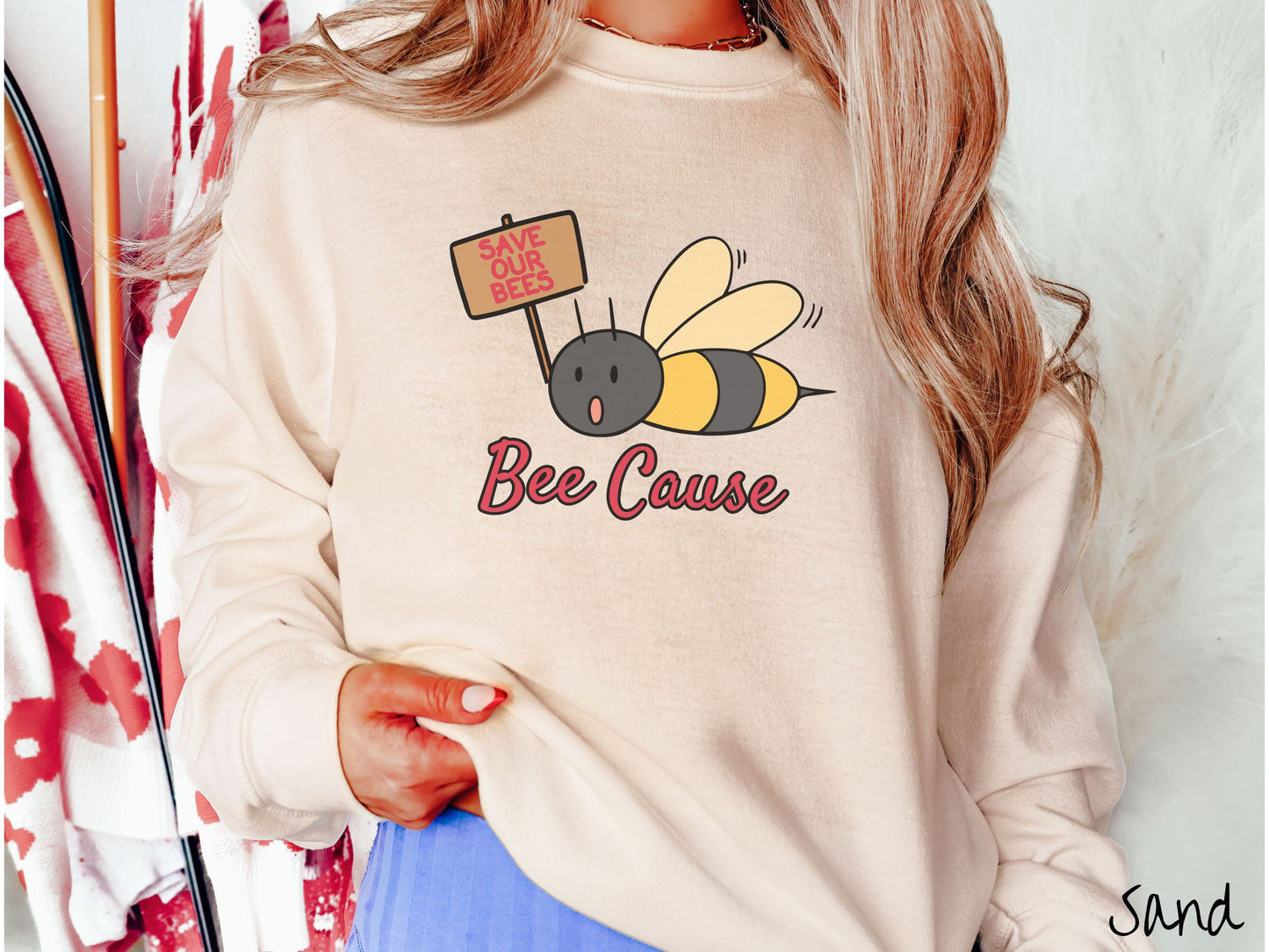 A woman wearing a cute, vintage sand colored comfy sweatshirt with the text Bee Cause in red, cursive font. Above that is a black and yellow honey bee speaking with its mouth open and holding a sign that reads Save Our Bees in red font.