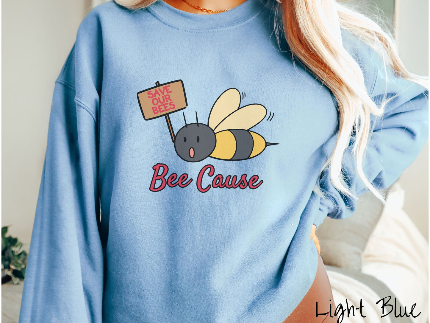 A woman wearing a cute, vintage light blue colored comfy sweatshirt with the text Bee Cause in red, cursive font. Above that is a black and yellow honey bee speaking with its mouth open and holding a sign that reads Save Our Bees in red font.