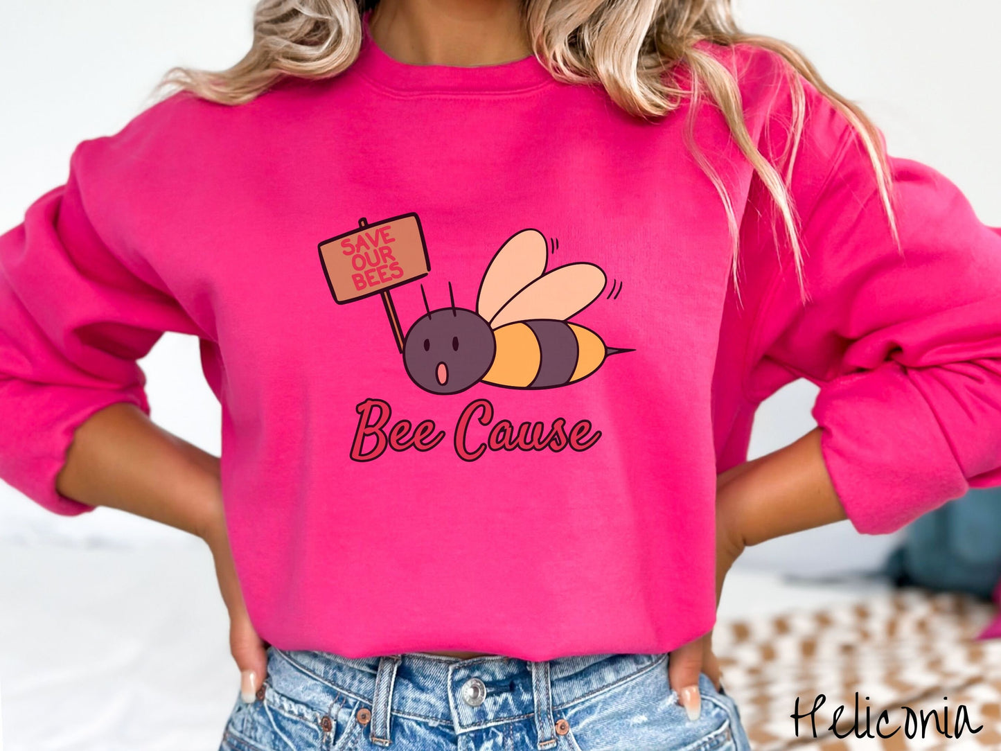 A woman wearing a cute, vintage heliconia colored comfy sweatshirt with the text Bee Cause in red, cursive font. Above that is a black and yellow honey bee speaking with its mouth open and holding a sign that reads Save Our Bees in red font.