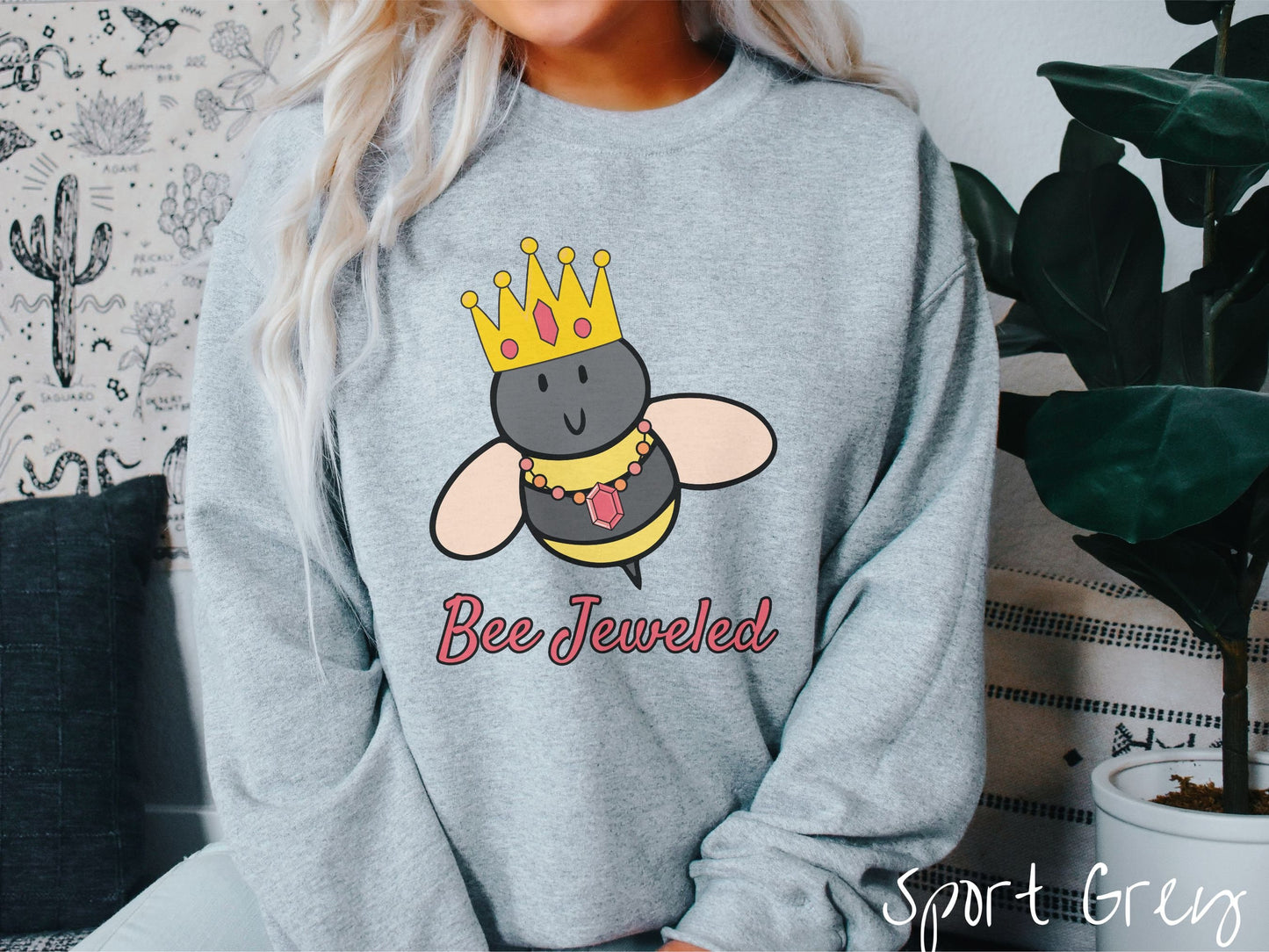 A woman wearing a cute, vintage sport grey colored comfy sweatshirt with the text Bee Jeweled in pink, cursive font. Above that is a black and yellow honey bee wearing a red, bejeweled golden crown, and a golden necklace with a large, red jewel.