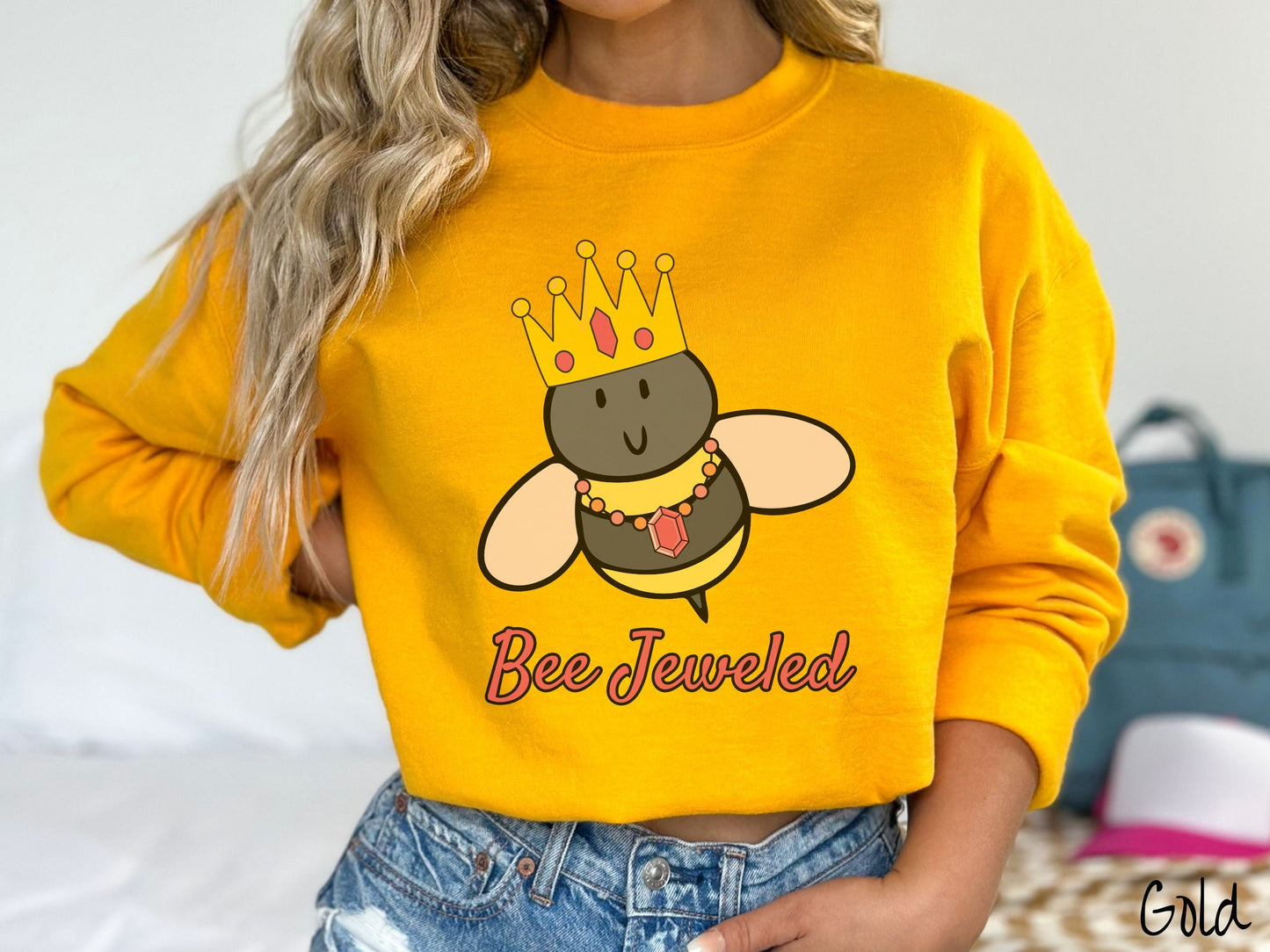 A woman wearing a cute, vintage gold colored comfy sweatshirt with the text Bee Jeweled in pink, cursive font. Above that is a black and yellow honey bee wearing a red, bejeweled golden crown, and a golden necklace with a large, red jewel.