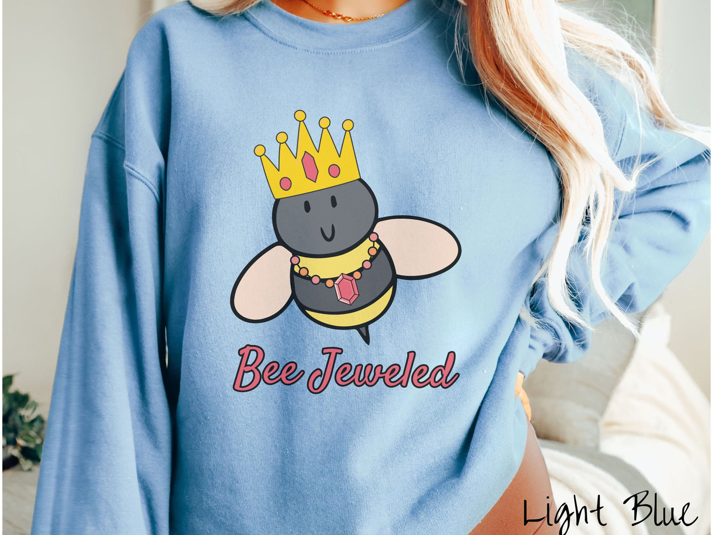 A woman wearing a cute, vintage light blue colored comfy sweatshirt with the text Bee Jeweled in pink, cursive font. Above that is a black and yellow honey bee wearing a red, bejeweled golden crown, and a golden necklace with a large, red jewel.