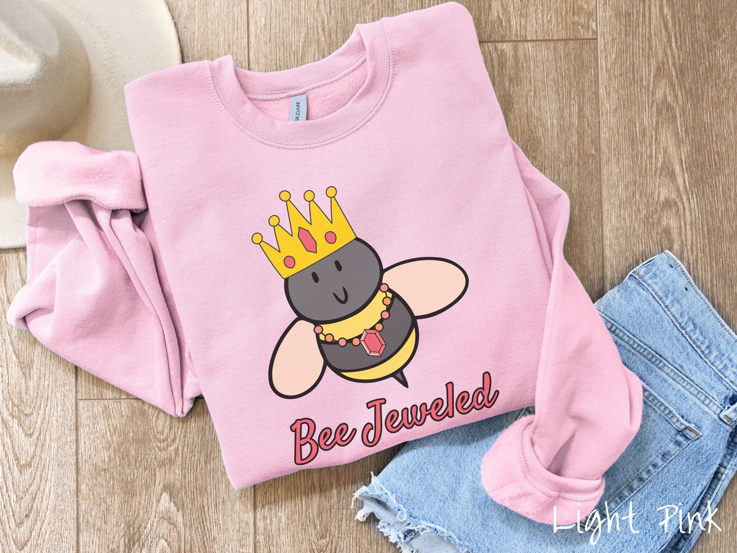 A cute, vintage light pink colored comfy sweatshirt with the text Bee Jeweled in pink, cursive font. Above that is a black and yellow honey bee wearing a red, bejeweled golden crown, and a golden necklace with a large, red jewel.