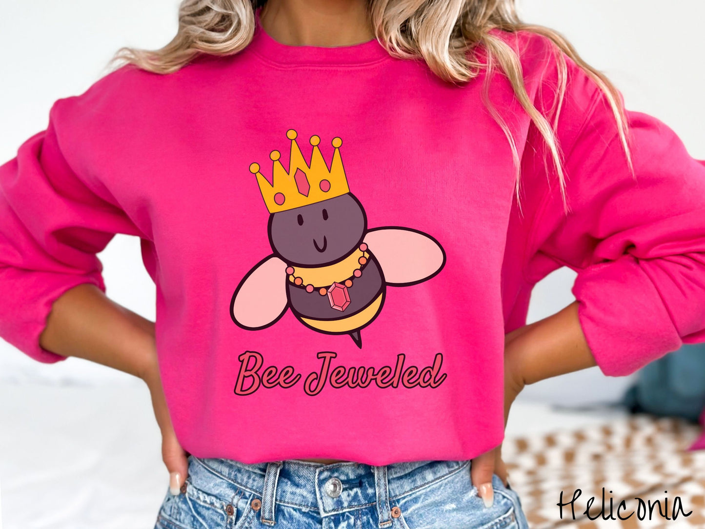 A woman wearing a cute, vintage heliconia colored comfy sweatshirt with the text Bee Jeweled in pink, cursive font. Above that is a black and yellow honey bee wearing a red, bejeweled golden crown, and a golden necklace with a large, red jewel.
