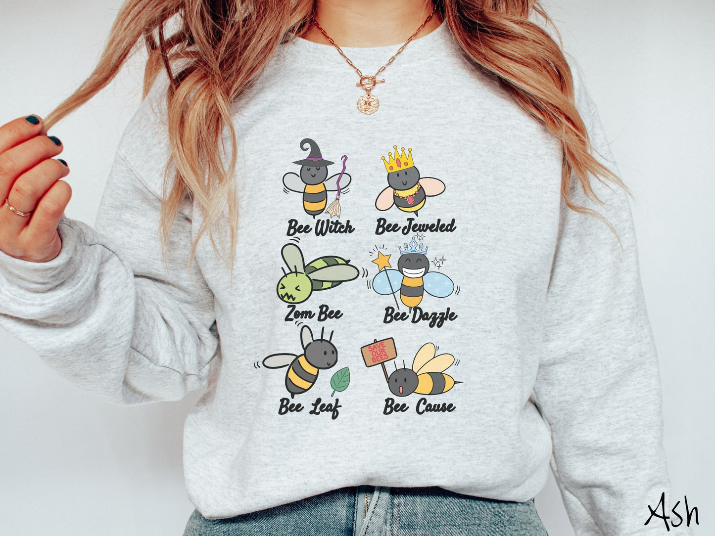 A woman wearing a cute, vintage ash colored comfy sweatshirt that has six puns that play on honey bees accompanied with bee drawings. The puns are Bee Witch, Bee Jeweled, Zom Bee, Bee Dazzle, Bee Leaf, and Bee Cause.