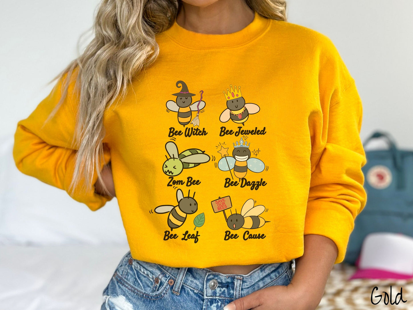 A woman wearing a cute, vintage gold colored comfy sweatshirt that has six puns that play on honey bees accompanied with bee drawings. The puns are Bee Witch, Bee Jeweled, Zom Bee, Bee Dazzle, Bee Leaf, and Bee Cause.