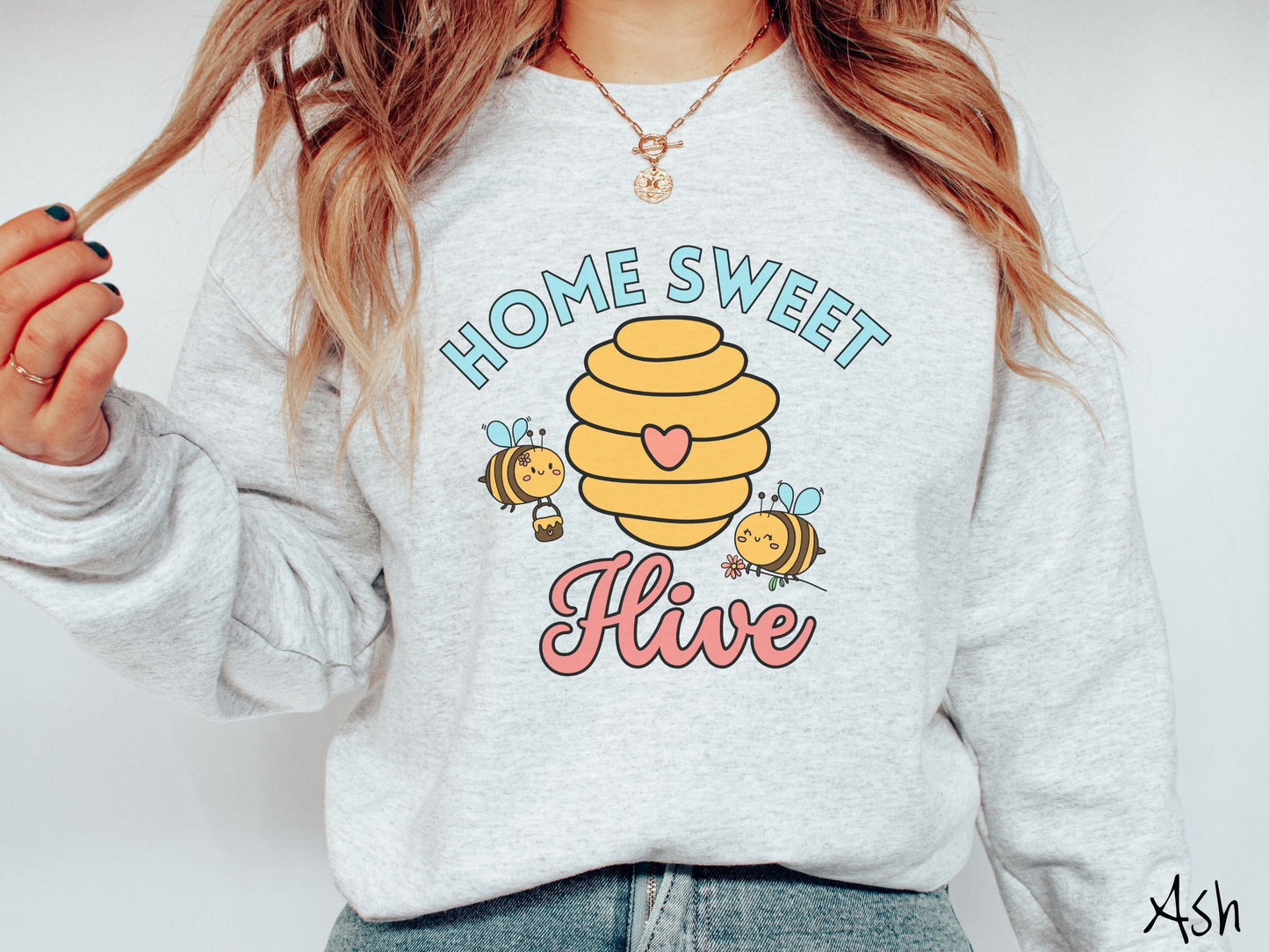 A woman wearing a cute, vintage ash colored comfy sweatshirt with the text Home Sweet Hive in cyan and red font. In between the words are a yellow bee hive with a red heart on it and smiling honey bees flying around holding flowers.