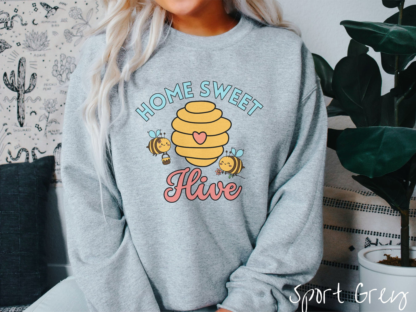 A woman wearing a cute, vintage sport grey colored comfy sweatshirt with the text Home Sweet Hive in cyan and red font. In between the words are a yellow bee hive with a red heart on it and smiling honey bees flying around holding flowers.