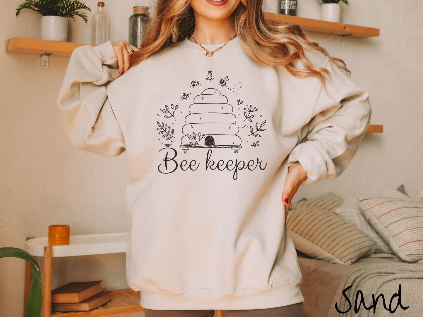 A woman wearing a cute, vintage sand colored comfy sweatshirt with the text Beekeeper across the front in black, cursive font. Above this is a black-outlined bee hive with bees buzzing around it and there are leaves in the air.