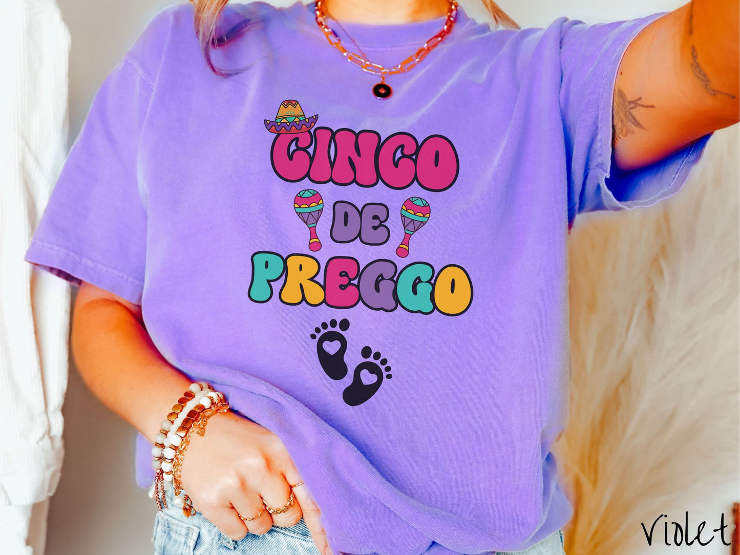 A woman wearing a cute, vintage violet colored Comfort Colors t-shirt with the text Cinco de Preggo in colorful font. A sombrero is hanging on top of the C in Cinco, colorful maracas around the word de, and two baby footprints with hearts inside.