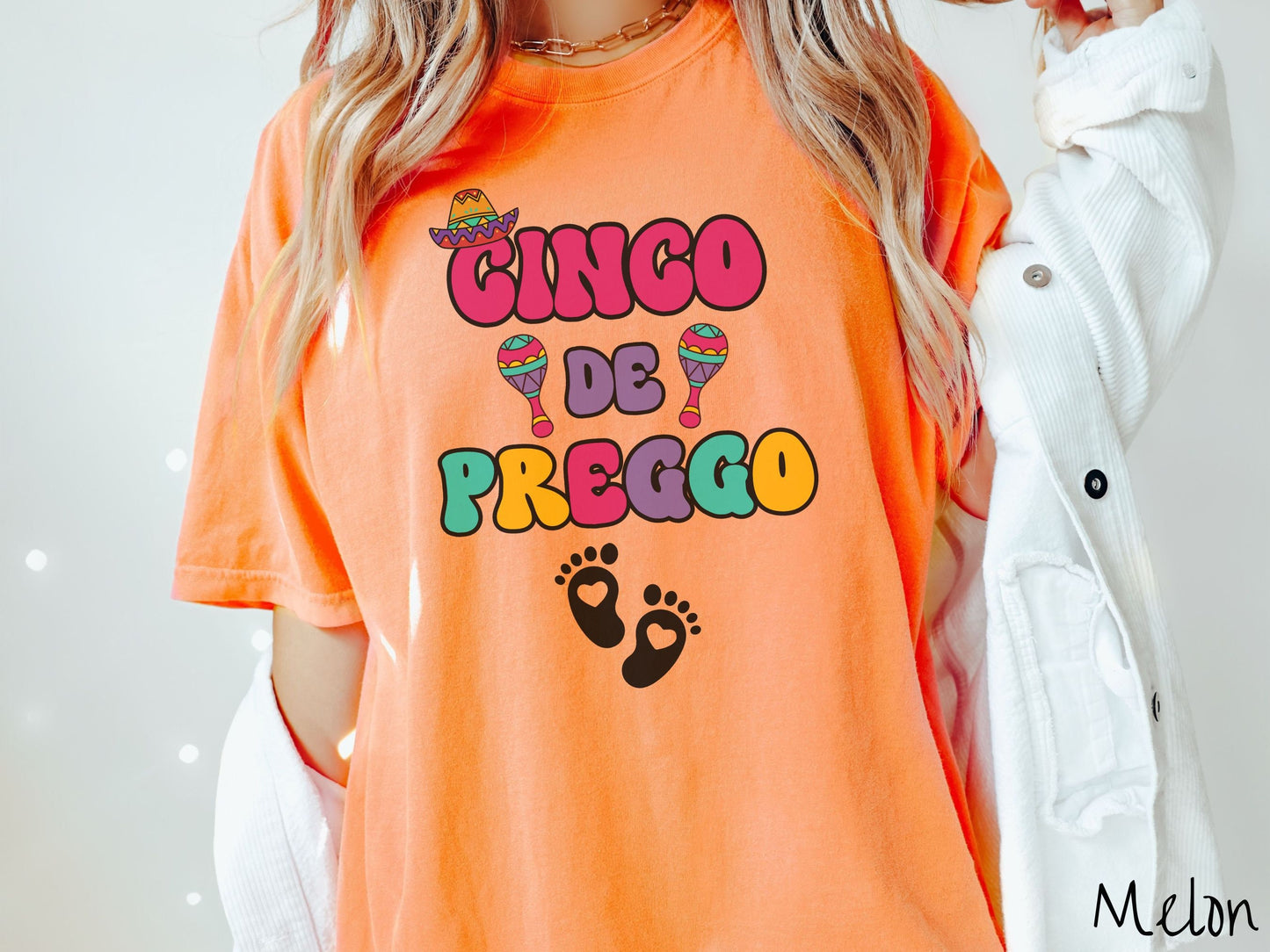 A woman wearing a cute, vintage melon colored Comfort Colors t-shirt with the text Cinco de Preggo in colorful font. A sombrero is hanging on top of the C in Cinco, colorful maracas around the word de, and two baby footprints with hearts inside.