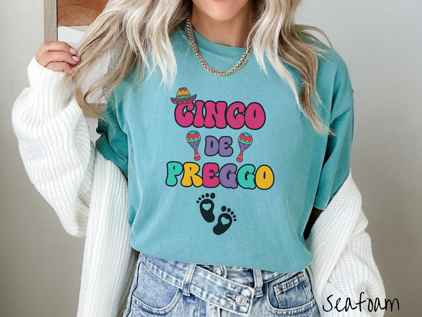 A woman wearing a cute, vintage seafoam colored Comfort Colors t-shirt with the text Cinco de Preggo in colorful font. A sombrero is hanging on top of the C in Cinco, colorful maracas around the word de, and two baby footprints with hearts inside.