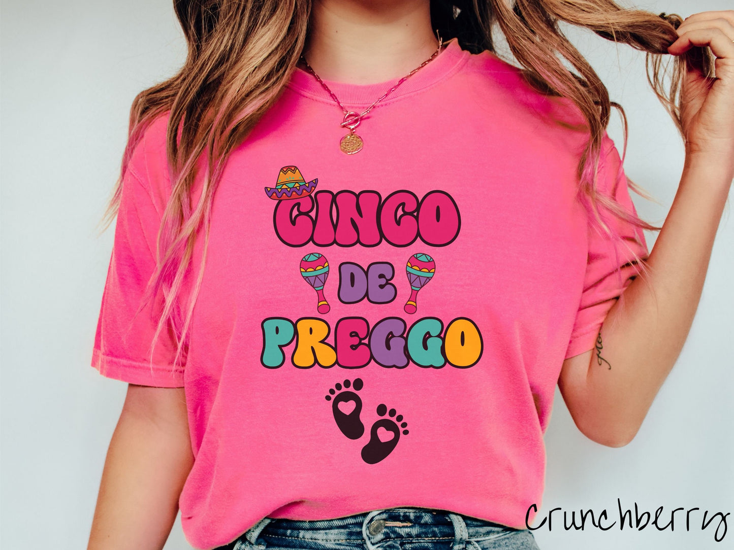 A woman wearing a cute, vintage crunchberry colored Comfort Colors t-shirt with text Cinco de Preggo in colorful font. A sombrero is hanging on top of the C in Cinco, colorful maracas around the word de, and two baby footprints with hearts inside.