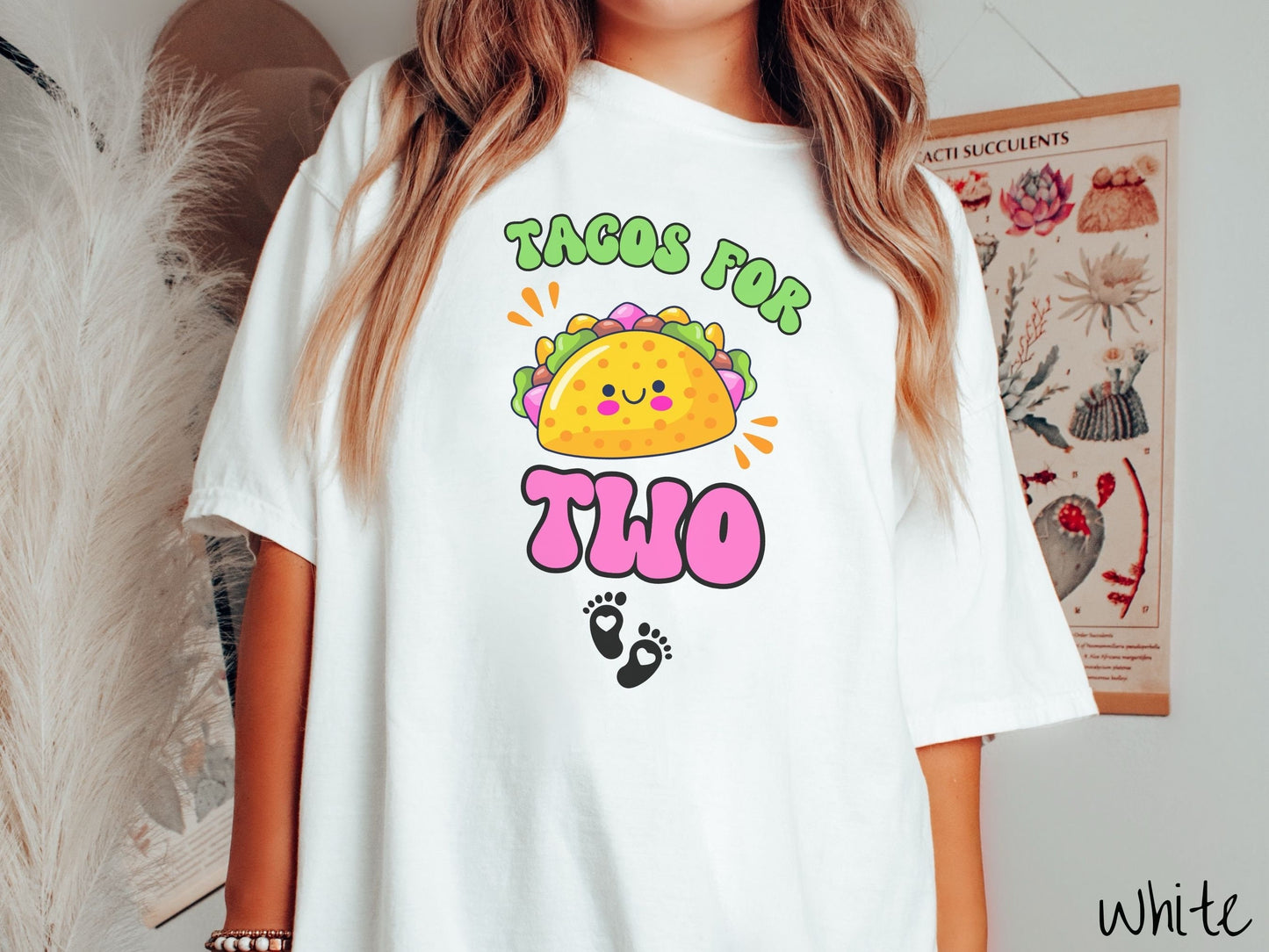 A woman wearing a cute, vintage white colored Comfort Colors t-shirt with text Tacos for Two in green and pink font. In between the font is a smiling taco with colorful fillings and below are two baby footprints with hearts inside.