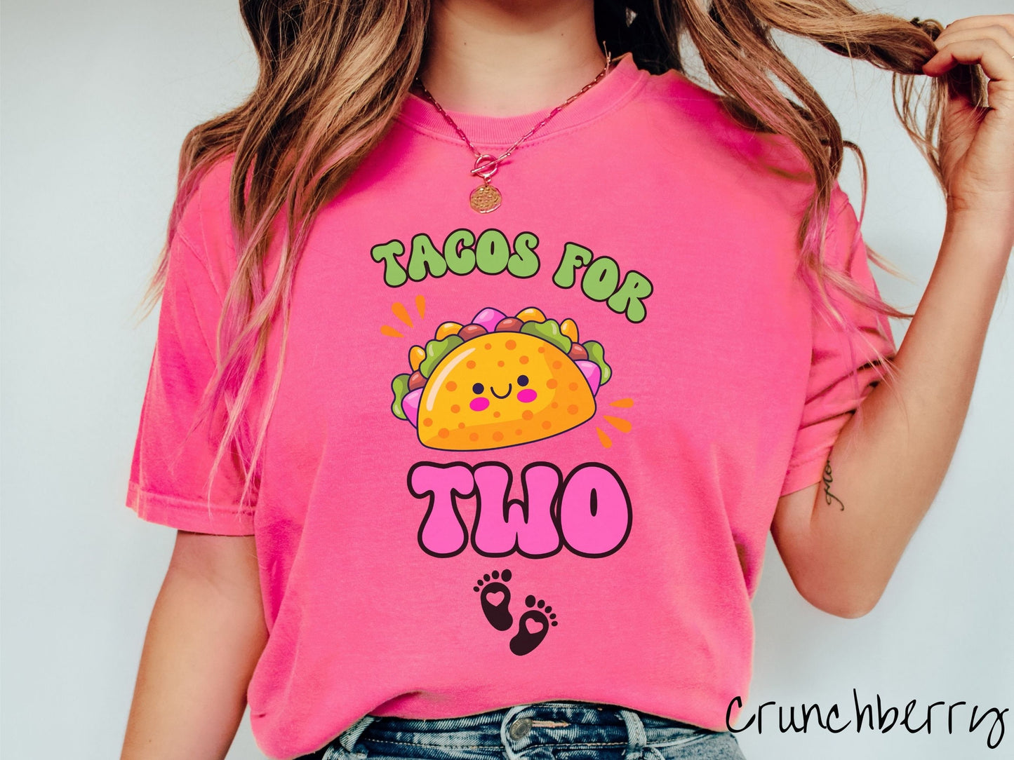 A woman wearing a cute, vintage crunchberry colored Comfort Colors t-shirt with text Tacos for Two in green and pink font. In between the font is a smiling taco with colorful fillings and below are two baby footprints with hearts inside.
