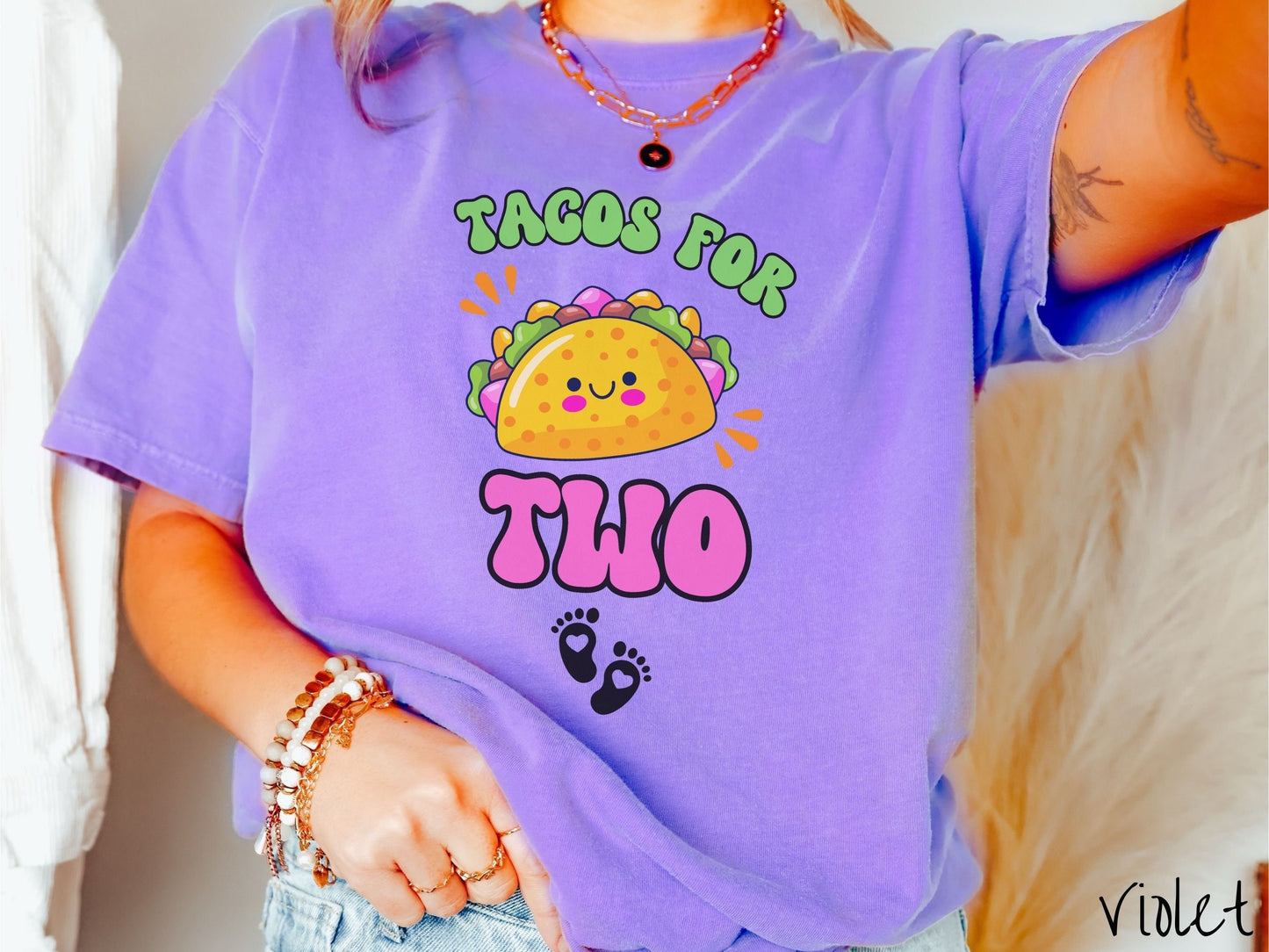 A woman wearing a cute, vintage violet colored Comfort Colors t-shirt with text Tacos for Two in green and pink font. In between the font is a smiling taco with colorful fillings and below are two baby footprints with hearts inside.