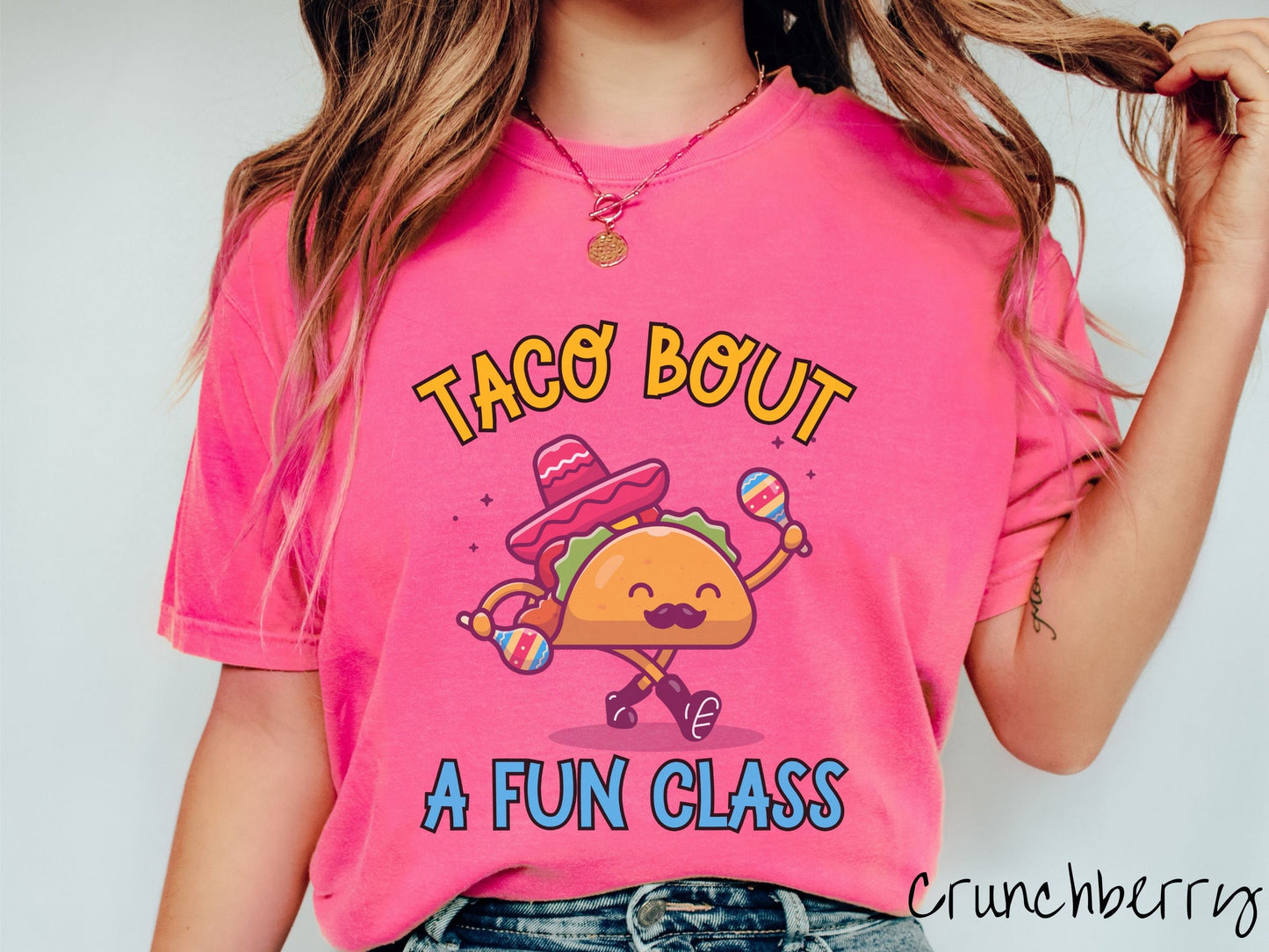 A woman wearing a cute, vintage crunchberry colored Comfort Colors t-shirt with text Taco Bout a Fun Class in yellow and blue font. In between the text is a smiling taco with a black mustache shaking colorful tambourines and wearing a red sombrero.