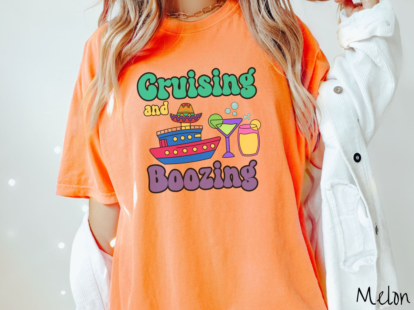 A woman wearing a cute, vintage melon colored Comfort Colors t-shirt with text Cruising and Boozing in green, yellow, and purple font. Between the text are a colorful cruise ship wearing a sombrero and two cocktail glasses with margaritas and limes.