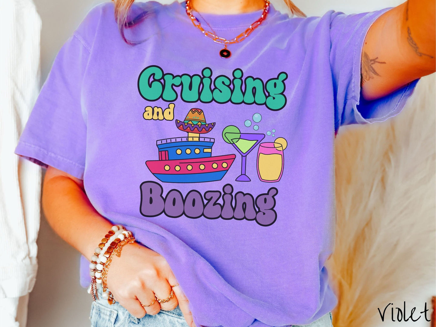 A woman wearing a cute, vintage violet colored Comfort Colors t-shirt with text Cruising and Boozing in green, yellow, and purple font. Between the text are a colorful cruise ship wearing a sombrero and two cocktail glasses with margaritas and limes.
