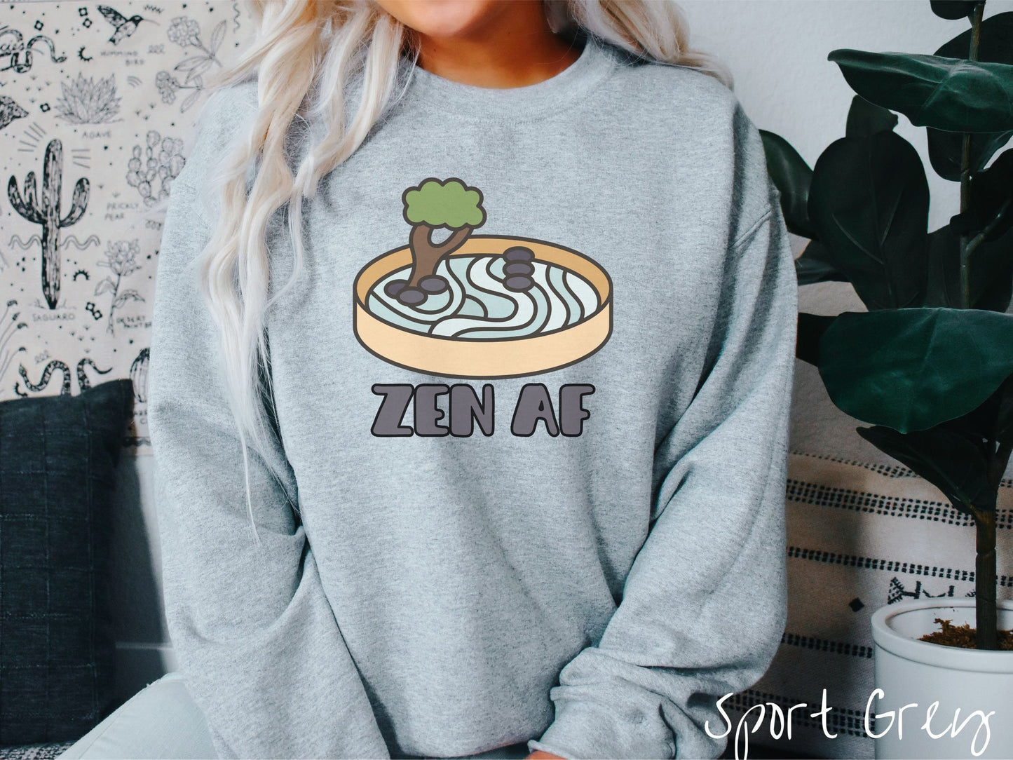 A woman wearing a cute, vintage sport grey colored comfy sweatshirt with the text Zen AF in gray font across the font. Above this is a zen garden in a yellow container with gray and light green sand, some black pebbles, and a green tree.