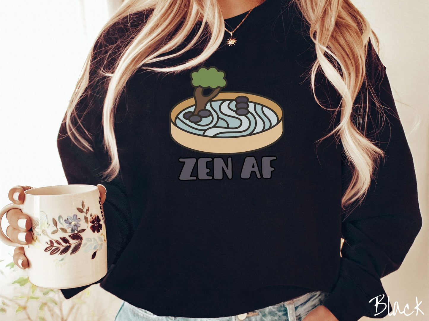 A woman wearing a cute, vintage black colored comfy sweatshirt with the text Zen AF in gray font across the font. Above this is a zen garden in a yellow container with gray and light green sand, some black pebbles, and a green tree.