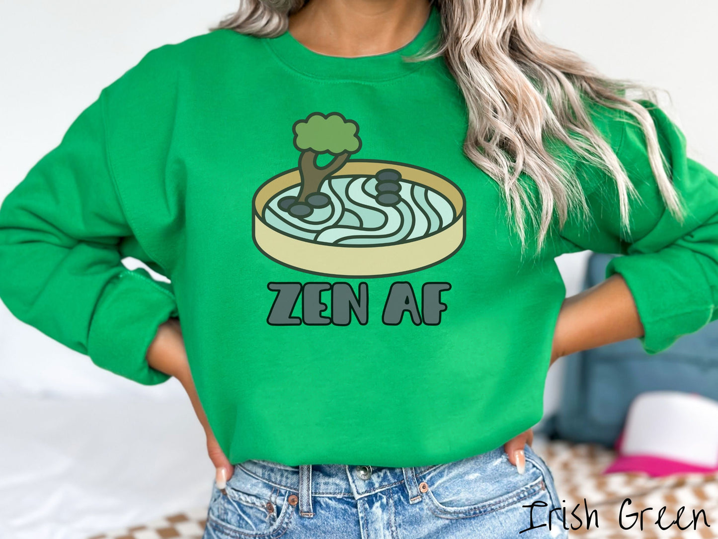 A woman wearing a cute, vintage Irish green colored comfy sweatshirt with the text Zen AF in gray font across the font. Above this is a zen garden in a yellow container with gray and light green sand, some black pebbles, and a green tree.