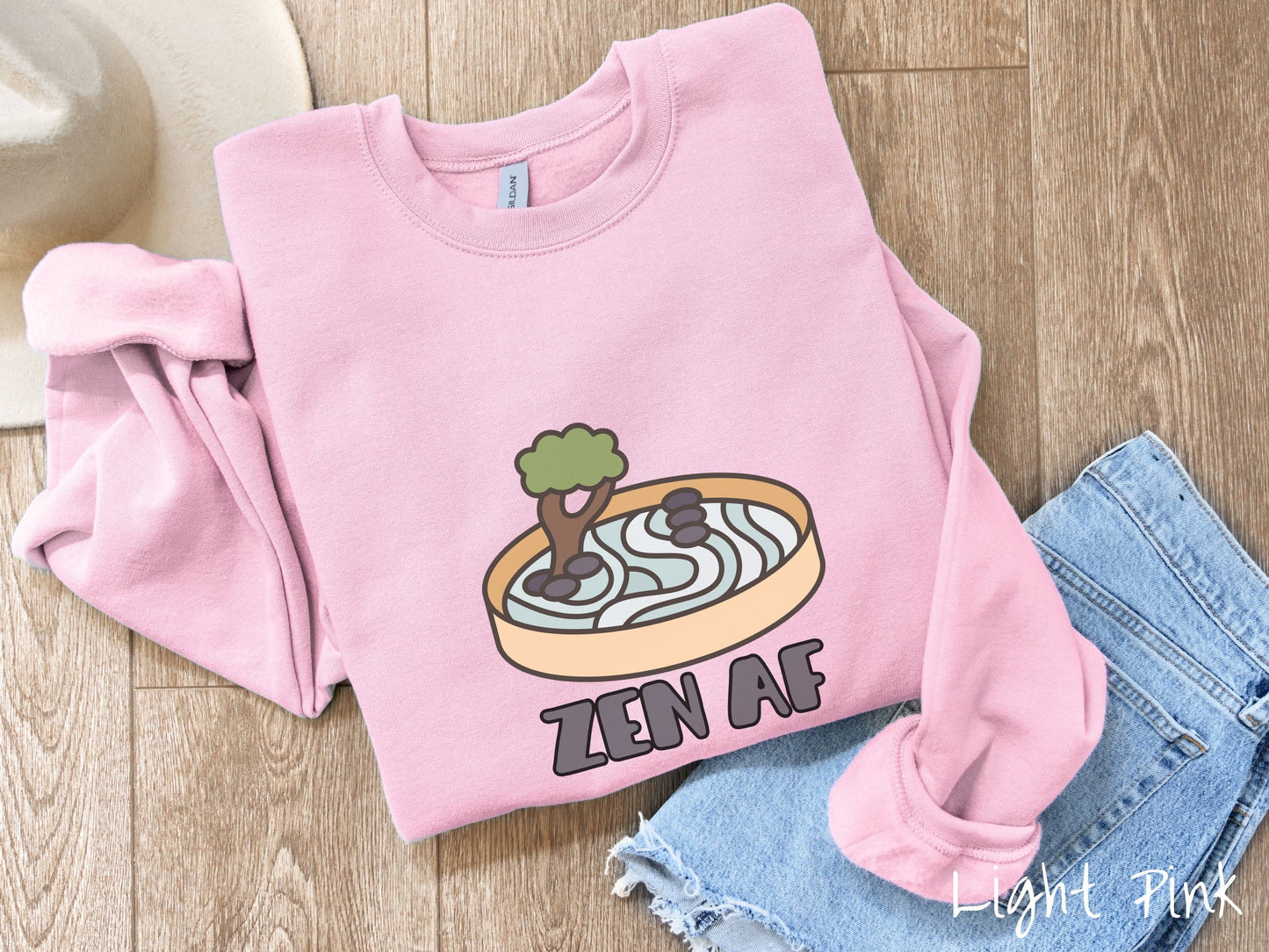 A cute, vintage light pink colored comfy sweatshirt with the text Zen AF in gray font across the font. Above this is a zen garden in a yellow container with gray and light green sand, some black pebbles, and a green tree.