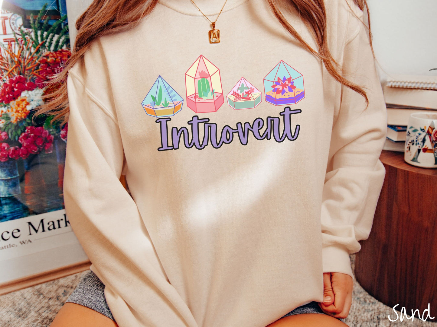 A woman wearing a cute, vintage sand colored comfy sweatshirt with the text Introvert in purple font across the front. Above the text are four colorful plant terrariums with colorful flowers and cacti inside.
