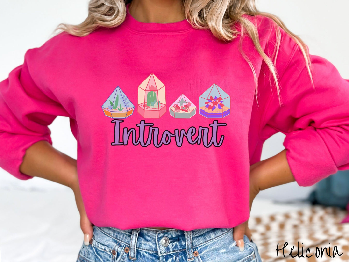 A woman wearing a cute, vintage heliconia colored comfy sweatshirt with the text Introvert in purple font across the front. Above the text are four colorful plant terrariums with colorful flowers and cacti inside.