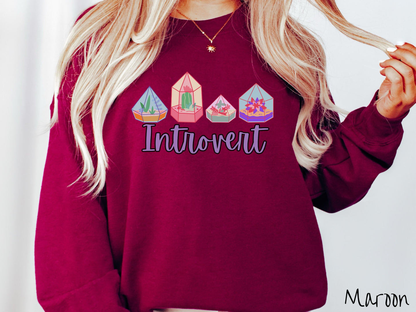 A woman wearing a cute, vintage maroon colored comfy sweatshirt with the text Introvert in purple font across the front. Above the text are four colorful plant terrariums with colorful flowers and cacti inside.
