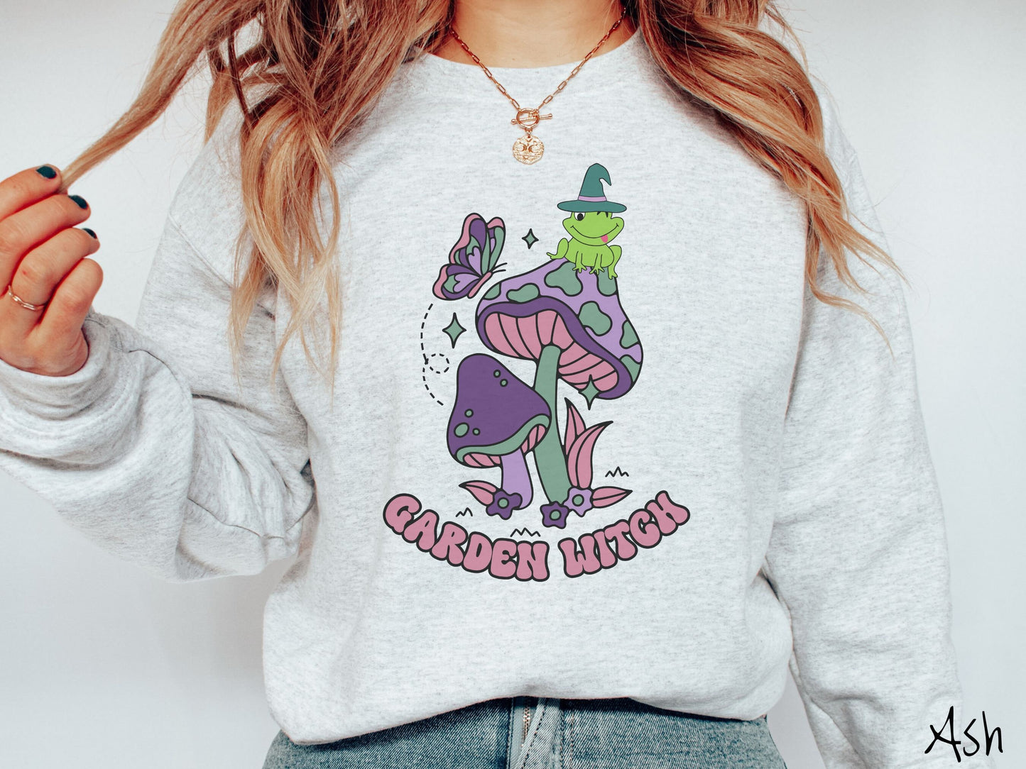 A woman wearing a cute, vintage ash colored comfy sweatshirt with the text Garden Witch in purple font. Above this text are two purple and green mushrooms with a green frog sitting atop one wearing a witch hat.