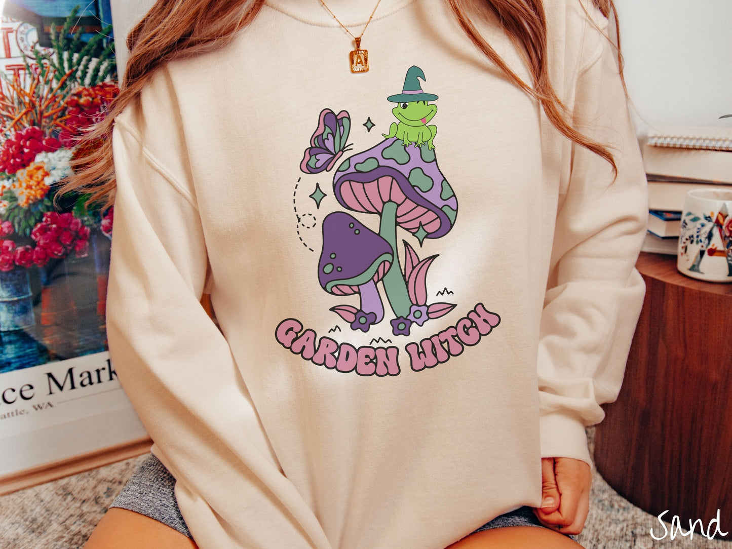 A woman wearing a cute, vintage sand colored comfy sweatshirt with the text Garden Witch in purple font. Above this text are two purple and green mushrooms with a green frog sitting atop one wearing a witch hat.