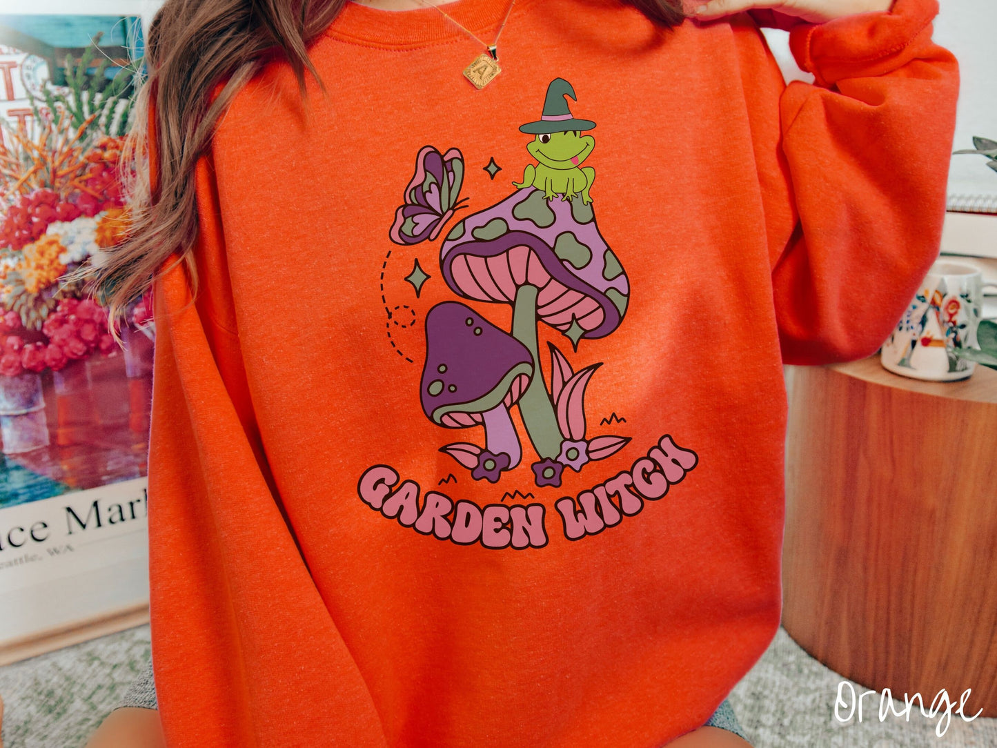 A woman wearing a cute, vintage orange colored comfy sweatshirt with the text Garden Witch in purple font. Above this text are two purple and green mushrooms with a green frog sitting atop one wearing a witch hat.