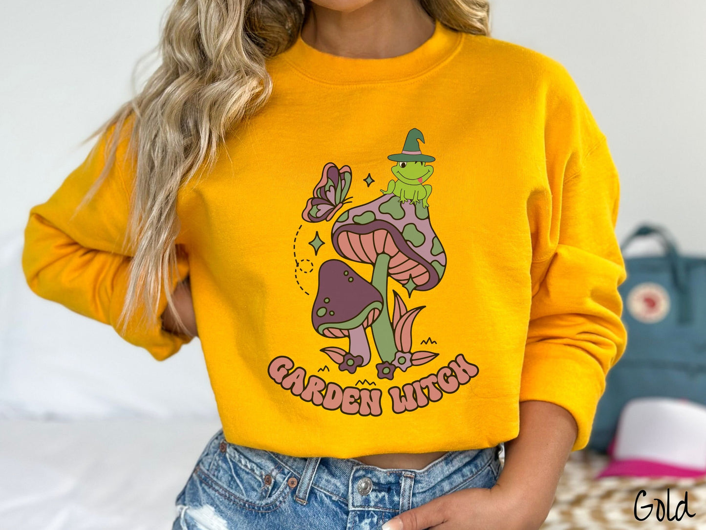 A woman wearing a cute, vintage gold colored comfy sweatshirt with the text Garden Witch in purple font. Above this text are two purple and green mushrooms with a green frog sitting atop one wearing a witch hat.