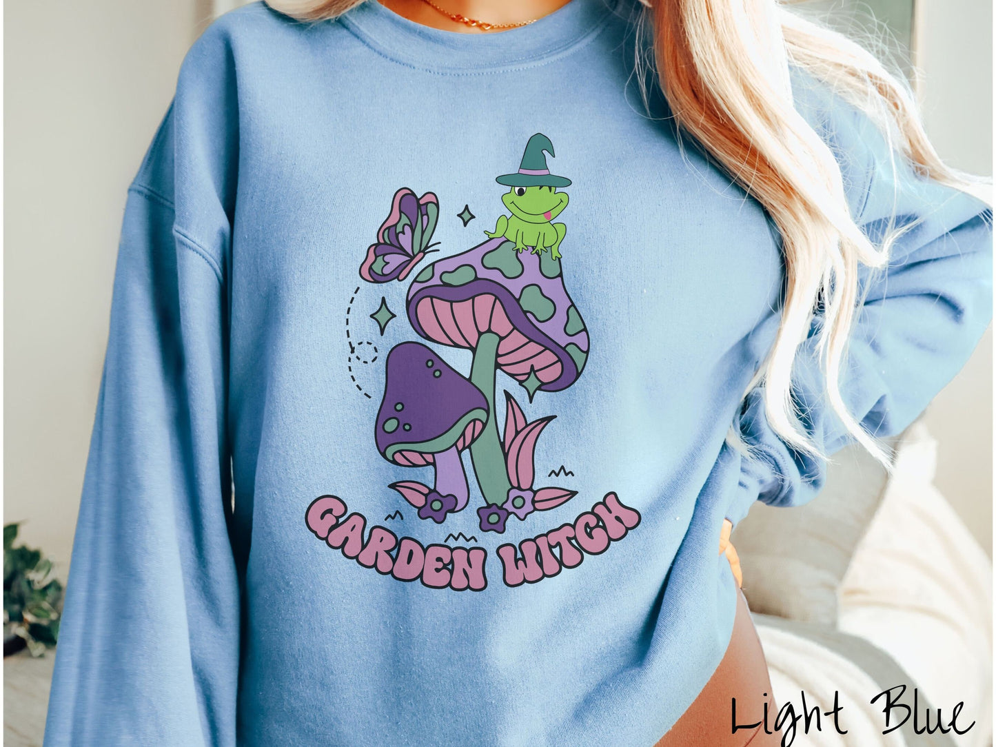 A woman wearing a cute, vintage light blue colored comfy sweatshirt with the text Garden Witch in purple font. Above this text are two purple and green mushrooms with a green frog sitting atop one wearing a witch hat.