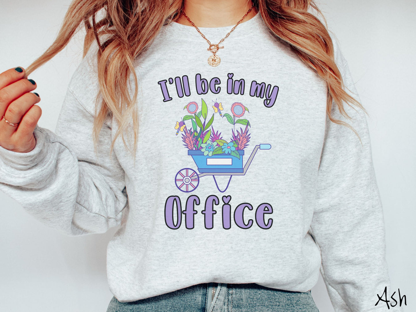 A woman wearing a cute, vintage ash colored comfy sweatshirt with the text I’ll Be in My Office in purple font across the front. In between the text is a blue wheelbarrow with a pink wheel stacked full of colorful plants and flowers.