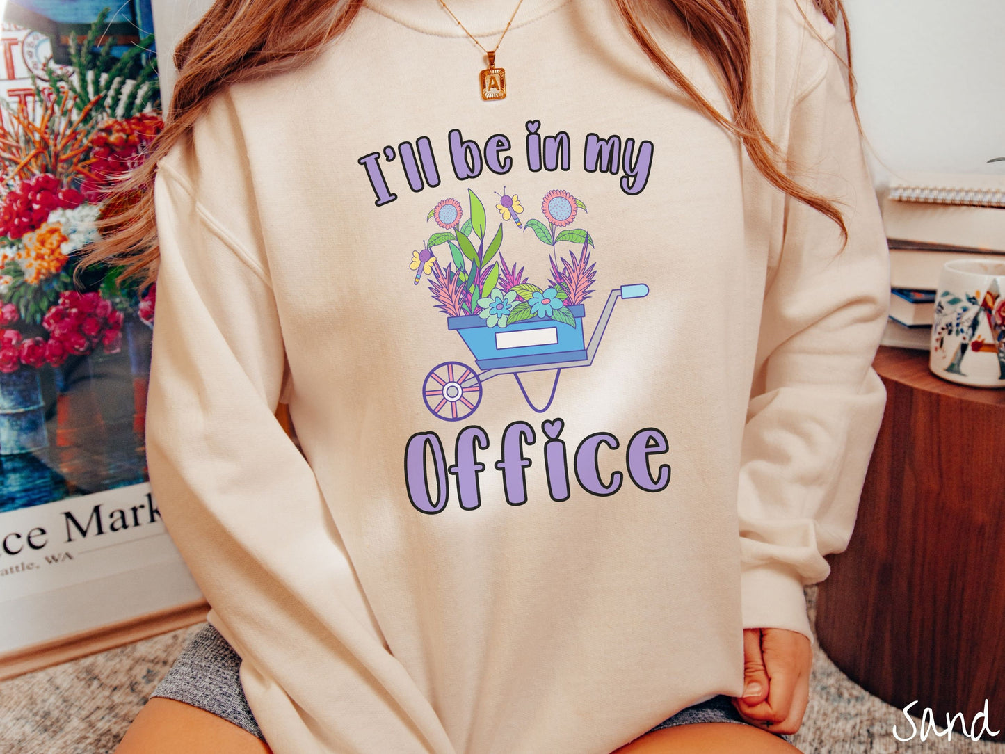 A woman wearing a cute, vintage sand colored comfy sweatshirt with the text I’ll Be in My Office in purple font across the front. In between the text is a blue wheelbarrow with a pink wheel stacked full of colorful plants and flowers.