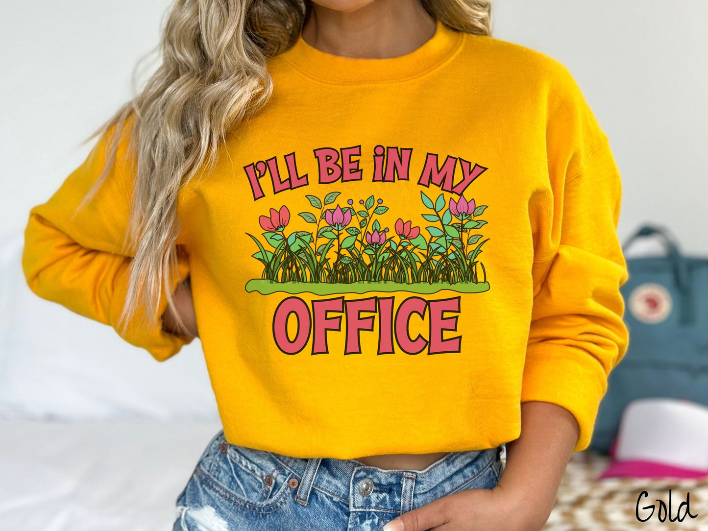 A woman wearing a cute, vintage gold colored comfy sweatshirt with the text I’ll Be in my Office in red font across the front. In between the text is a garden of red and pink flowers, green plants, and lush green grass.