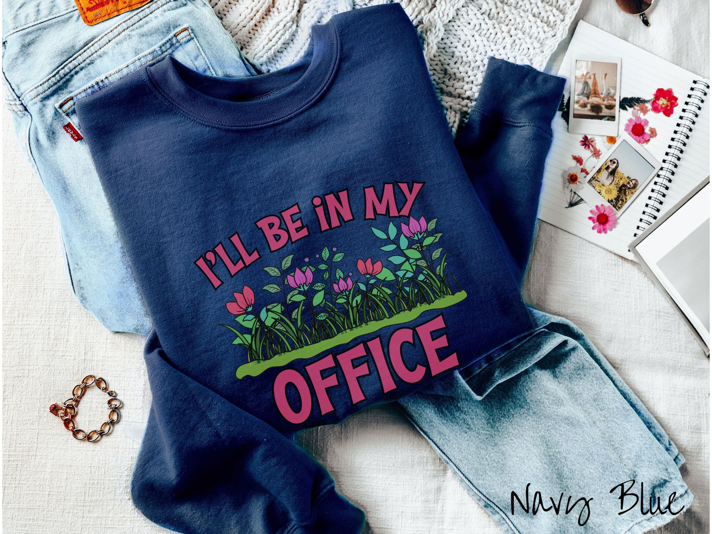 A cute, vintage navy blue colored comfy sweatshirt with the text I’ll Be in my Office in red font across the front. In between the text is a garden of red and pink flowers, green plants, and lush green grass.