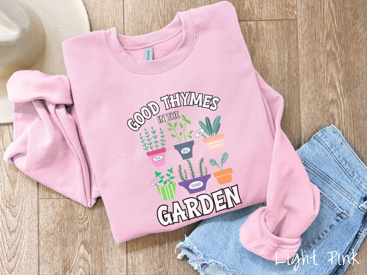 A cute, vintage light pink colored comfy sweatshirt with the text Good Thymes in the Garden in white font. In between the text are different potted vegetables in colorful pots like thyme, basil, sage, and mint.
