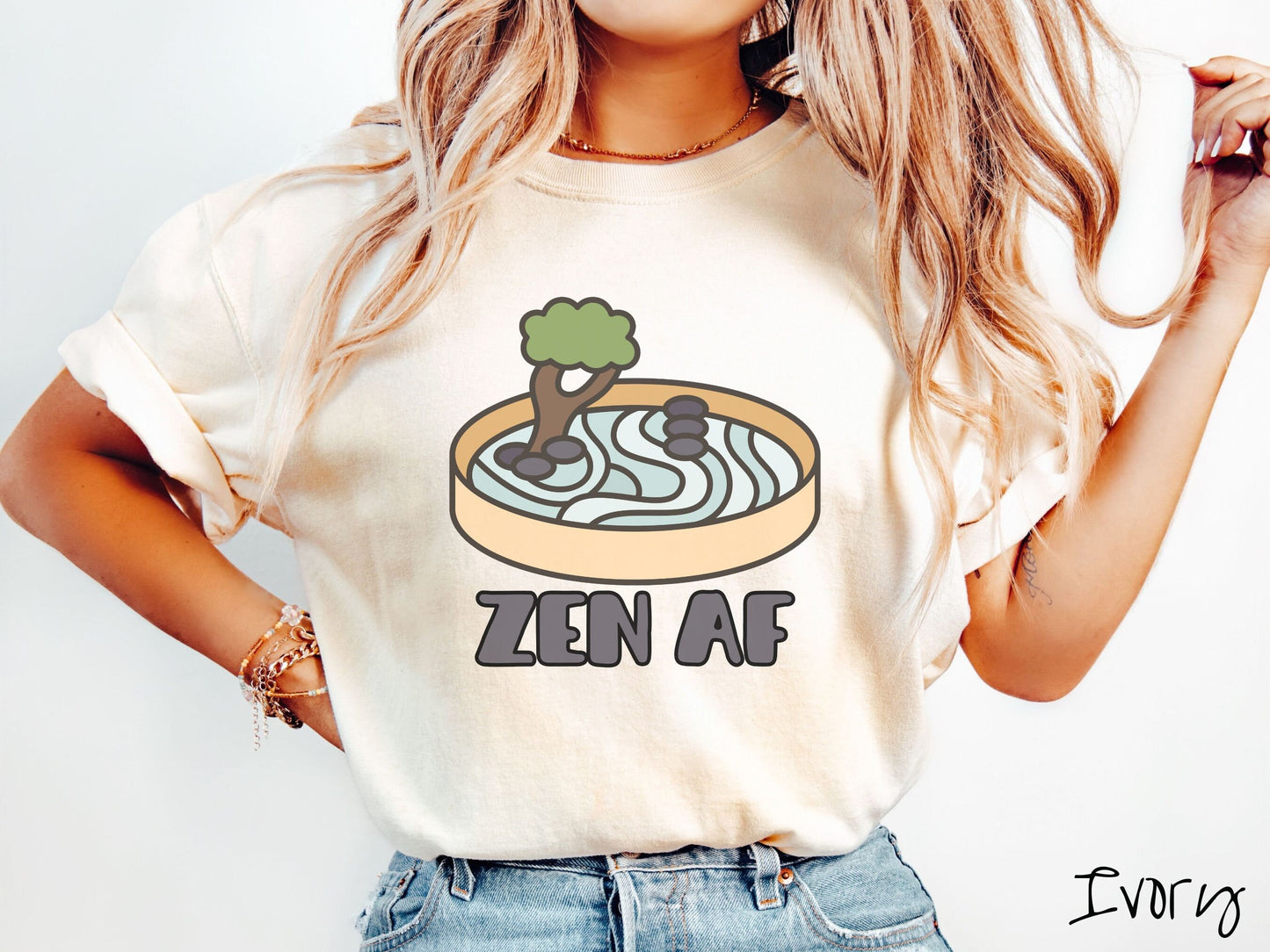 A woman wearing a cute, vintage ivory colored Comfort Colors t-shirt with the text Zen AF in gray font across the font. Above this is a zen garden in a yellow container with gray and light green sand, some black pebbles, and a green tree.