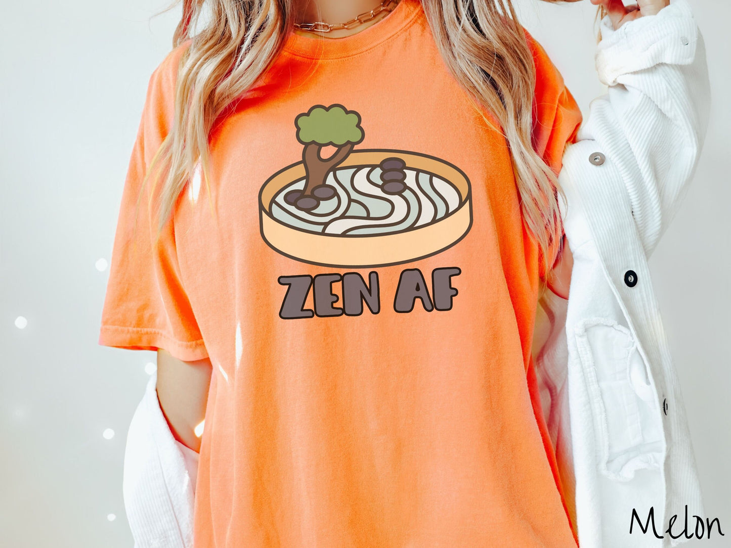 A woman wearing a cute, vintage melon colored Comfort Colors t-shirt with the text Zen AF in gray font across the font. Above this is a zen garden in a yellow container with gray and light green sand, some black pebbles, and a green tree.