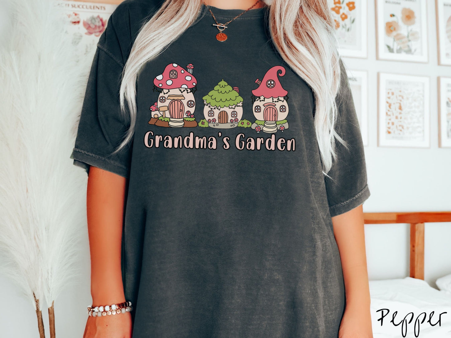 A woman wearing a cute, vintage pepper colored Comfort Colors t-shirt with the text Grandmas Garden in light pink font. Above the text are cute little cottages for gnomes made from light and dark pink mushrooms.