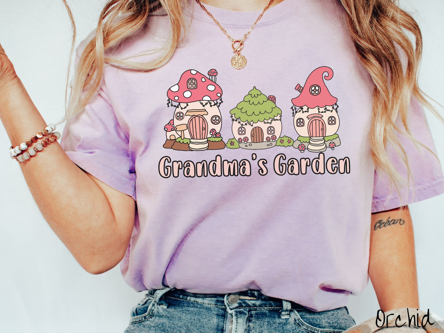 A woman wearing a cute, vintage orchid colored Comfort Colors t-shirt with the text Grandmas Garden in light pink font. Above the text are cute little cottages for gnomes made from light and dark pink mushrooms.