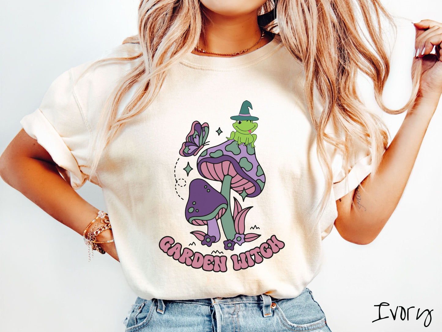 A woman wearing a cute, vintage ivory colored Comfort Colors t-shirt with the text Garden Witch in purple font. Above this text are two purple and green mushrooms with a green frog sitting atop one wearing a witch hat.