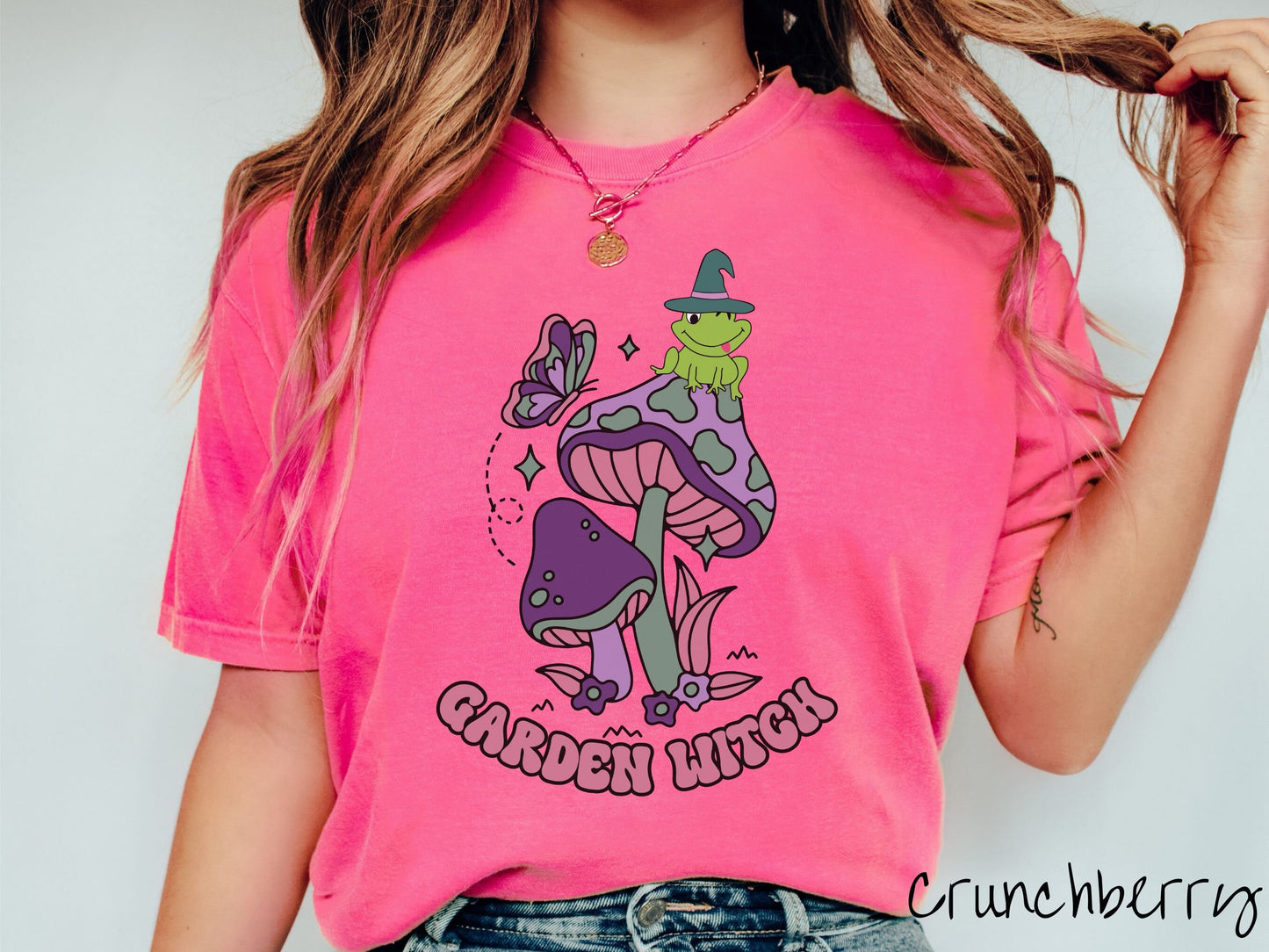 A woman wearing a cute, vintage crunchberry colored Comfort Colors t-shirt with the text Garden Witch in purple font. Above this text are two purple and green mushrooms with a green frog sitting atop one wearing a witch hat.