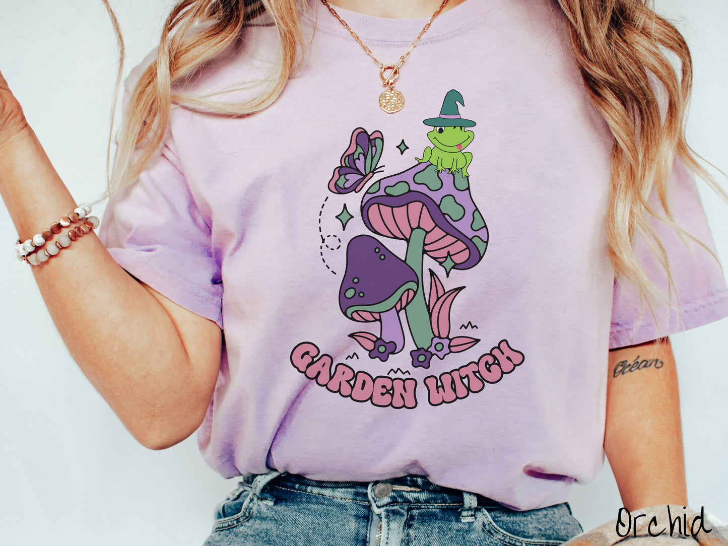 A woman wearing a cute, vintage orchid colored Comfort Colors t-shirt with the text Garden Witch in purple font. Above this text are two purple and green mushrooms with a green frog sitting atop one wearing a witch hat.