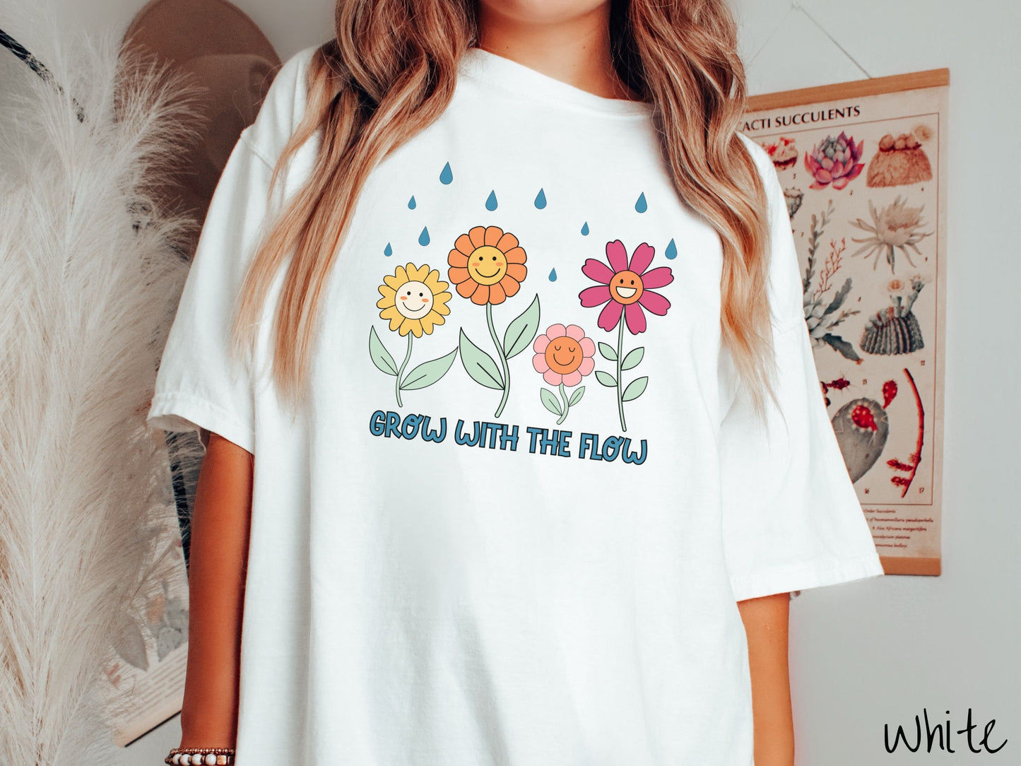 A woman wearing a cute, vintage white colored Comfort Colors t-shirt with the text Grow with the Flow in blue font across the bottom. Above the text are smiling yellow, orange, pink, and red flowers with raindrops falling in the background.