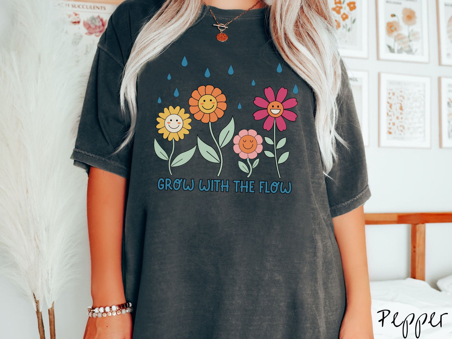 A woman wearing a cute, vintage pepper colored Comfort Colors t-shirt with the text Grow with the Flow in blue font across the bottom. Above the text are smiling yellow, orange, pink, and red flowers with raindrops falling in the background.