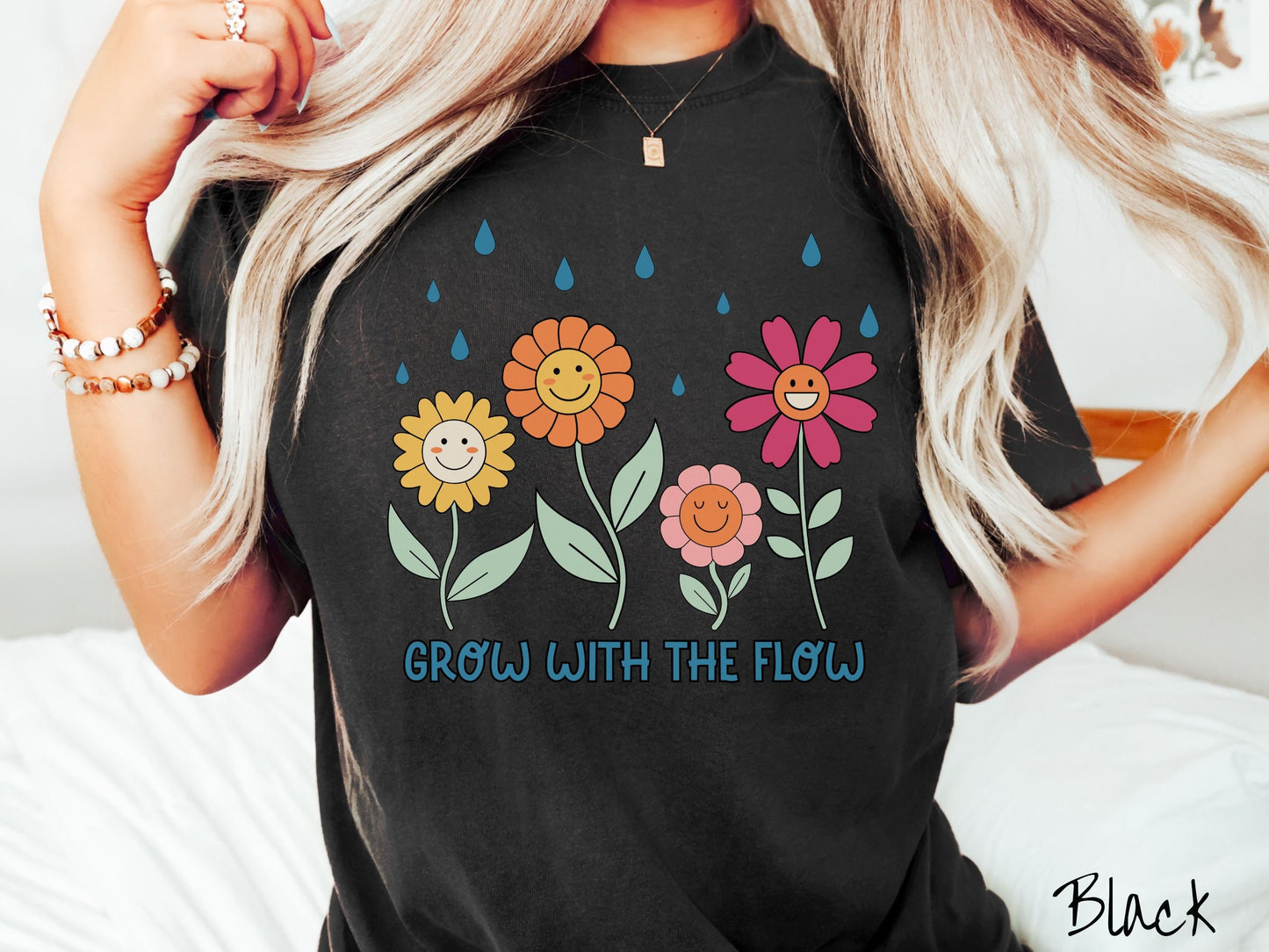 A woman wearing a cute, vintage black colored Comfort Colors t-shirt with the text Grow with the Flow in blue font across the bottom. Above the text are smiling yellow, orange, pink, and red flowers with raindrops falling in the background.